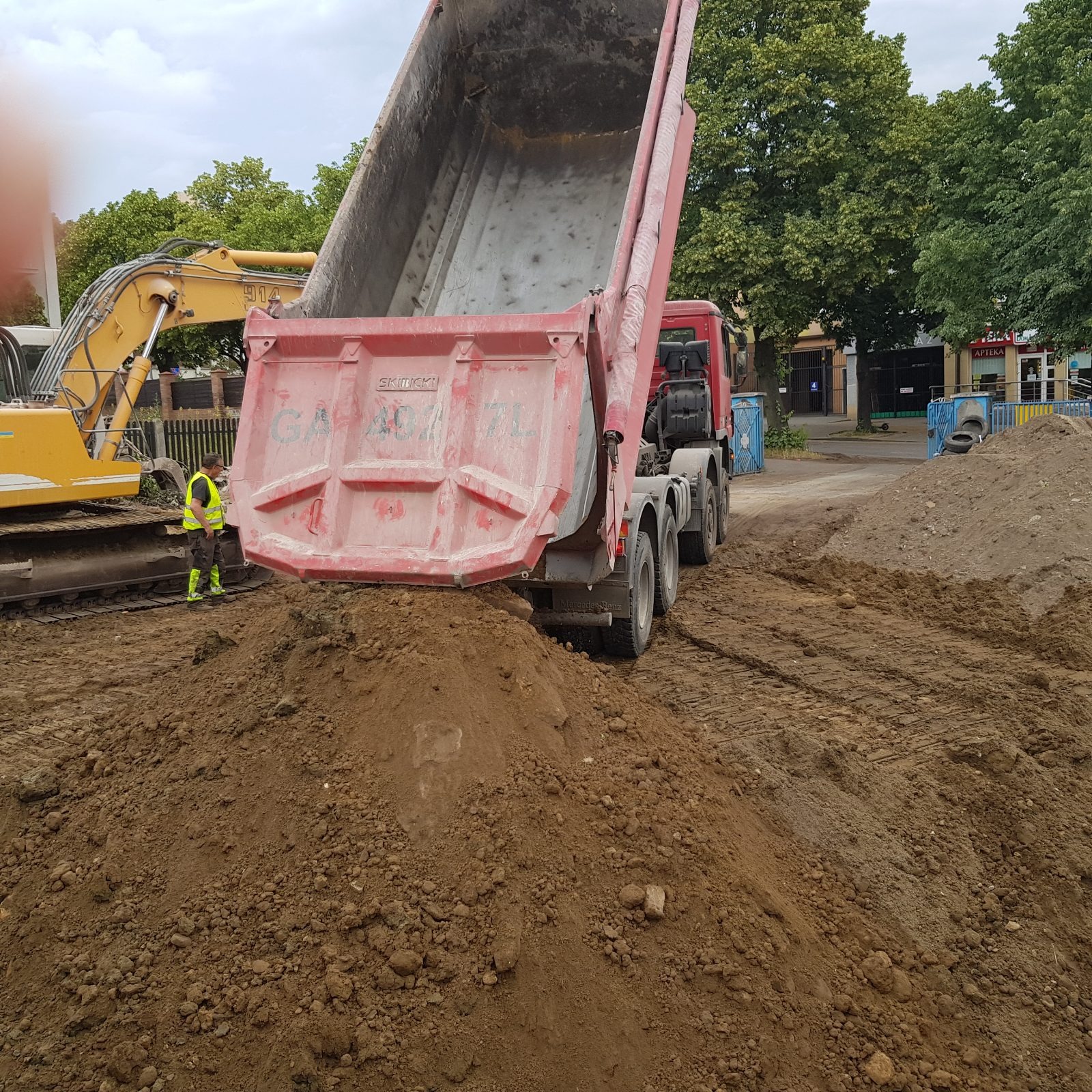 Ciężarówka z podniesioną skrzynią wysypuje ziemię na pryzmę. W tle żółta koparka i pracownik w kamizelce odblaskowej. Prace ziemne w otoczeniu miejskiej zabudowy.