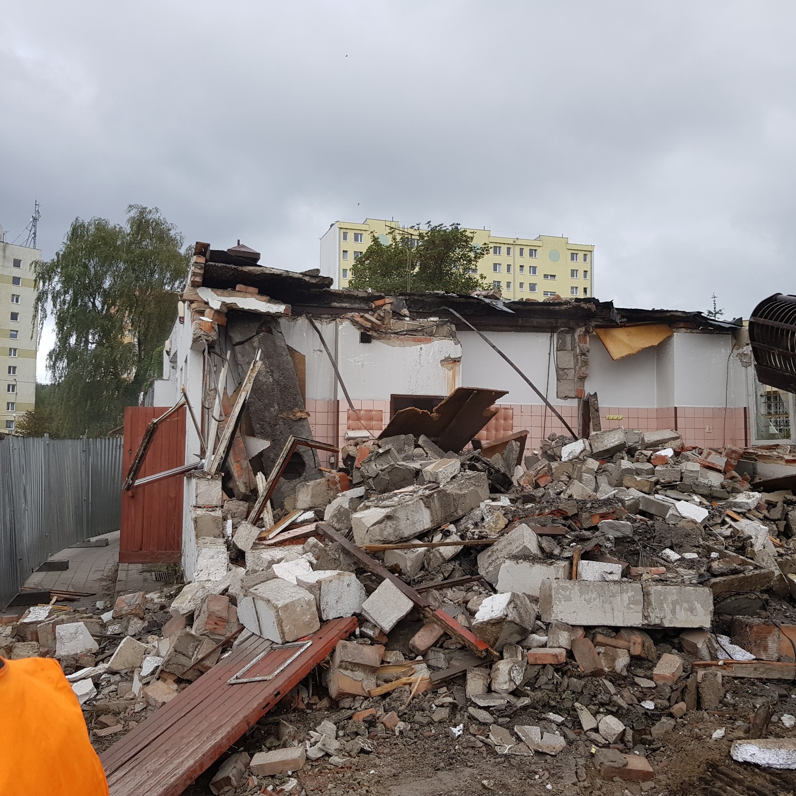 Zniszczony budynek po rozbiórce z widocznymi gruzami, fragmentami ścian i dachu, na tle zabudowy miejskiej i pochmurnego nieba. Widoczny fragment pomarańczowej odzieży roboczej.