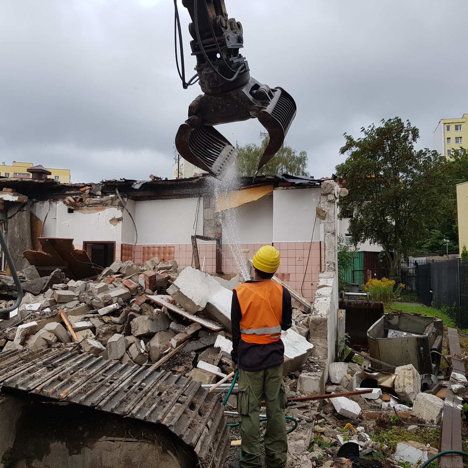 Rozbiórka budynku z użyciem maszyny z chwytakiem, pracownik w kasku spryskuje gruz wodą. Widoczne pozostałości ścian i elementy konstrukcyjne.