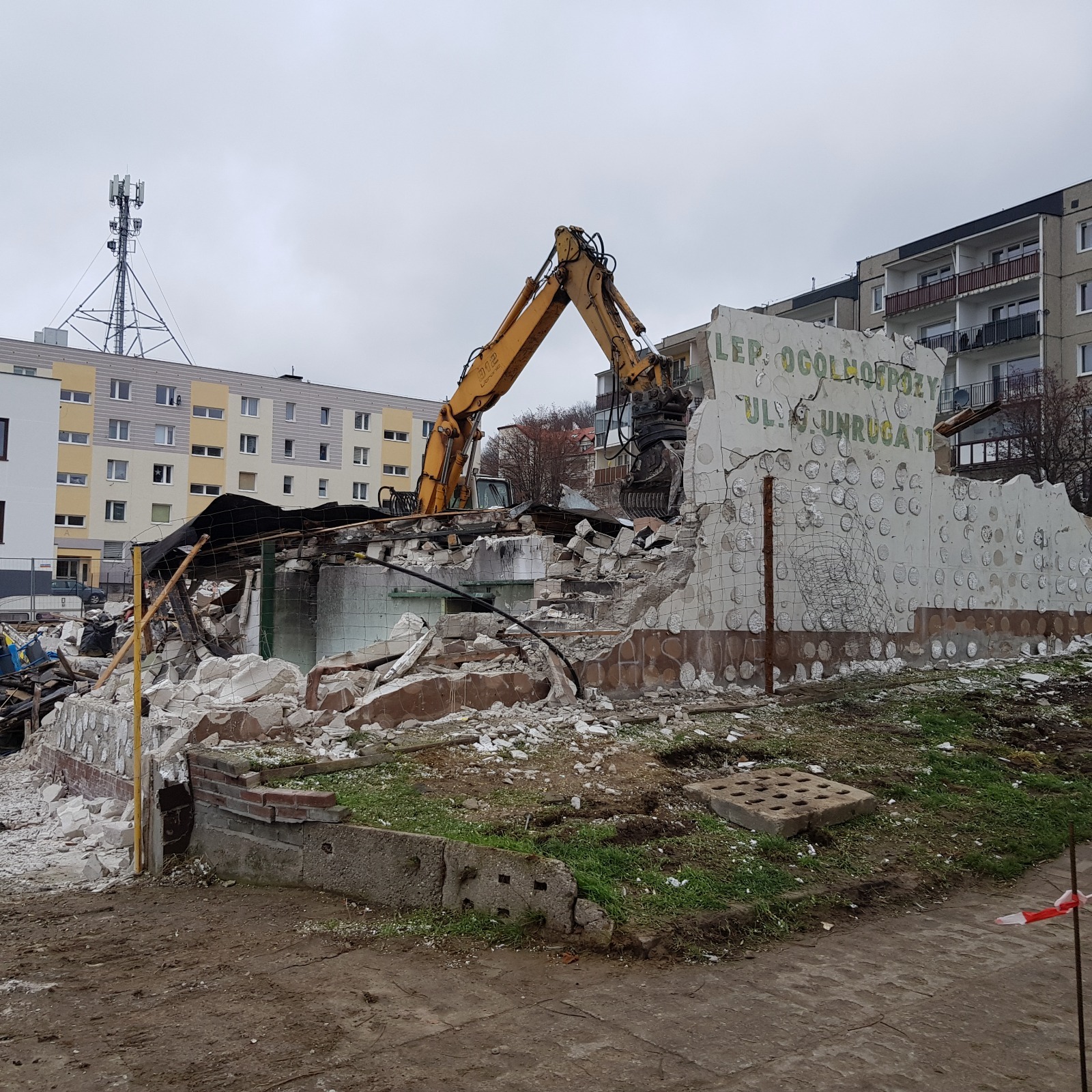 Żółta koparka z hydraulicznymi nożycami rozbiera resztki budynku z napisem 'LEP OGÓLNOSPŻY'. W tle bloki mieszkalne i wieża telekomunikacyjna.