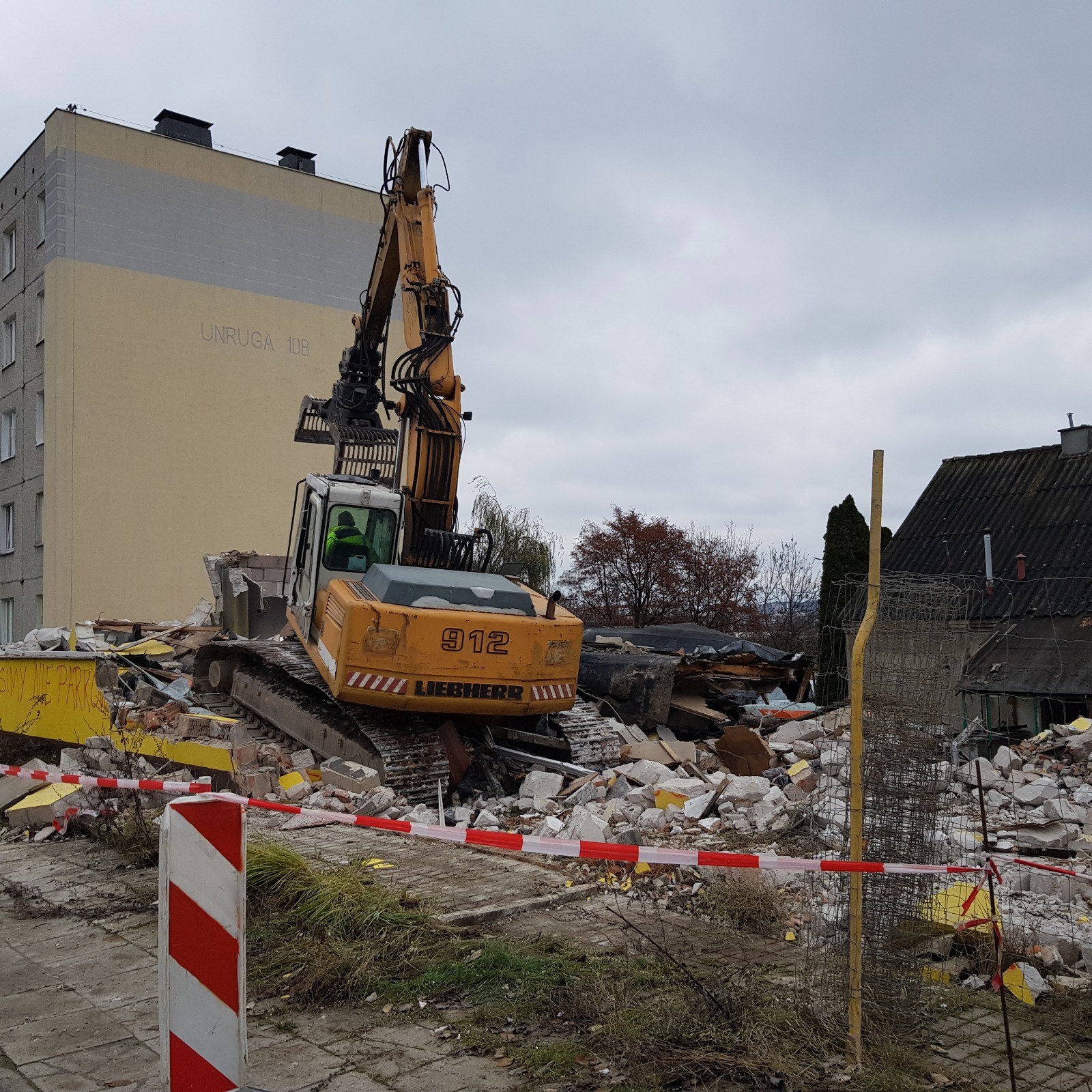 Żółta koparka Liebherr 912 na gruzowisku po rozbiórce budynku, z widocznym fragmentem bloku z napisem 'UNRUGA 108', otoczone taśmą ostrzegawczą.