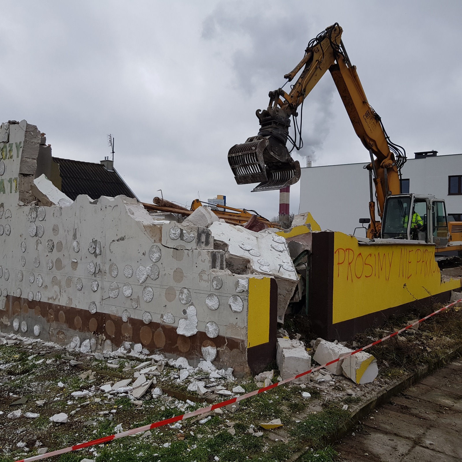 Żółta koparka z chwytakiem rozbiera fragment budynku z napisem 'Prosimy nie parkować' na tle pochmurnego nieba i komina. Gruz na ziemi.