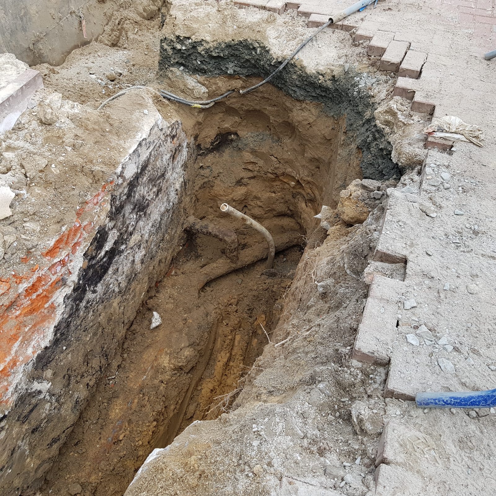 Głęboki wykop w ziemi z odsłoniętymi rurami i przewodami, ceglana ściana z elementami pomarańczowej farby, krawędź wyłożona cegłami, widoczne warstwy gruntu.