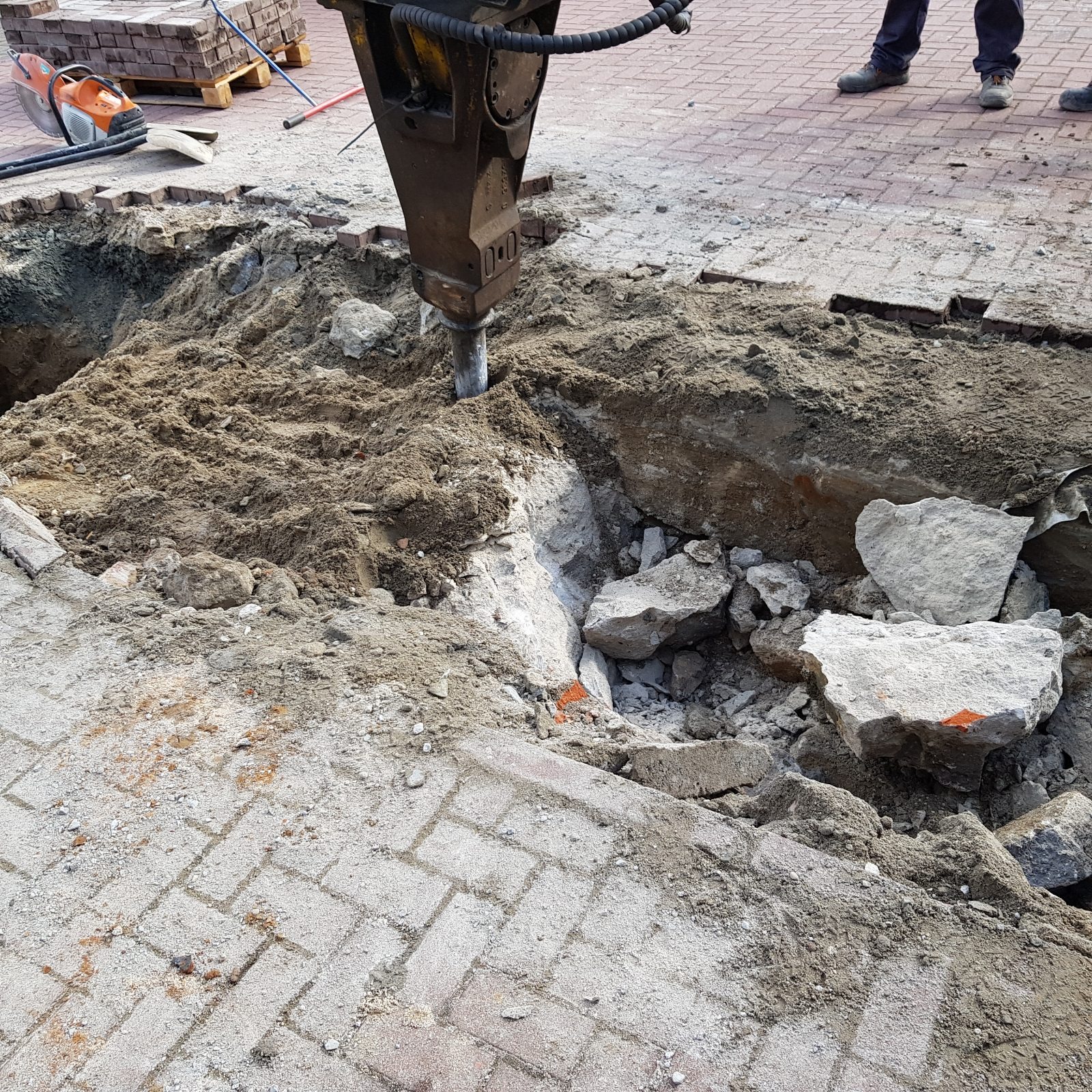 Hydrauliczny młot pneumatyczny kruszy betonową nawierzchnię. Widoczne gruz, piasek i fragmenty kostki brukowej. W tle paleta z kostką.