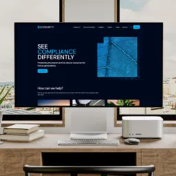 Ekran komputera stacjonarnego wyświetla stronę internetową Ecosurety z hasłem 'See Compliance Differently' i grafiką liścia na ciemnoniebieskim tle, umieszczony na drewnianym biurku z rośliną...