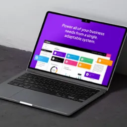 Laptop wyświetlający interfejs użytkownika aplikacji biznesowej z fioletowym tłem i kolorowymi elementami graficznymi, ustawiony pod kątem na szarym tle.