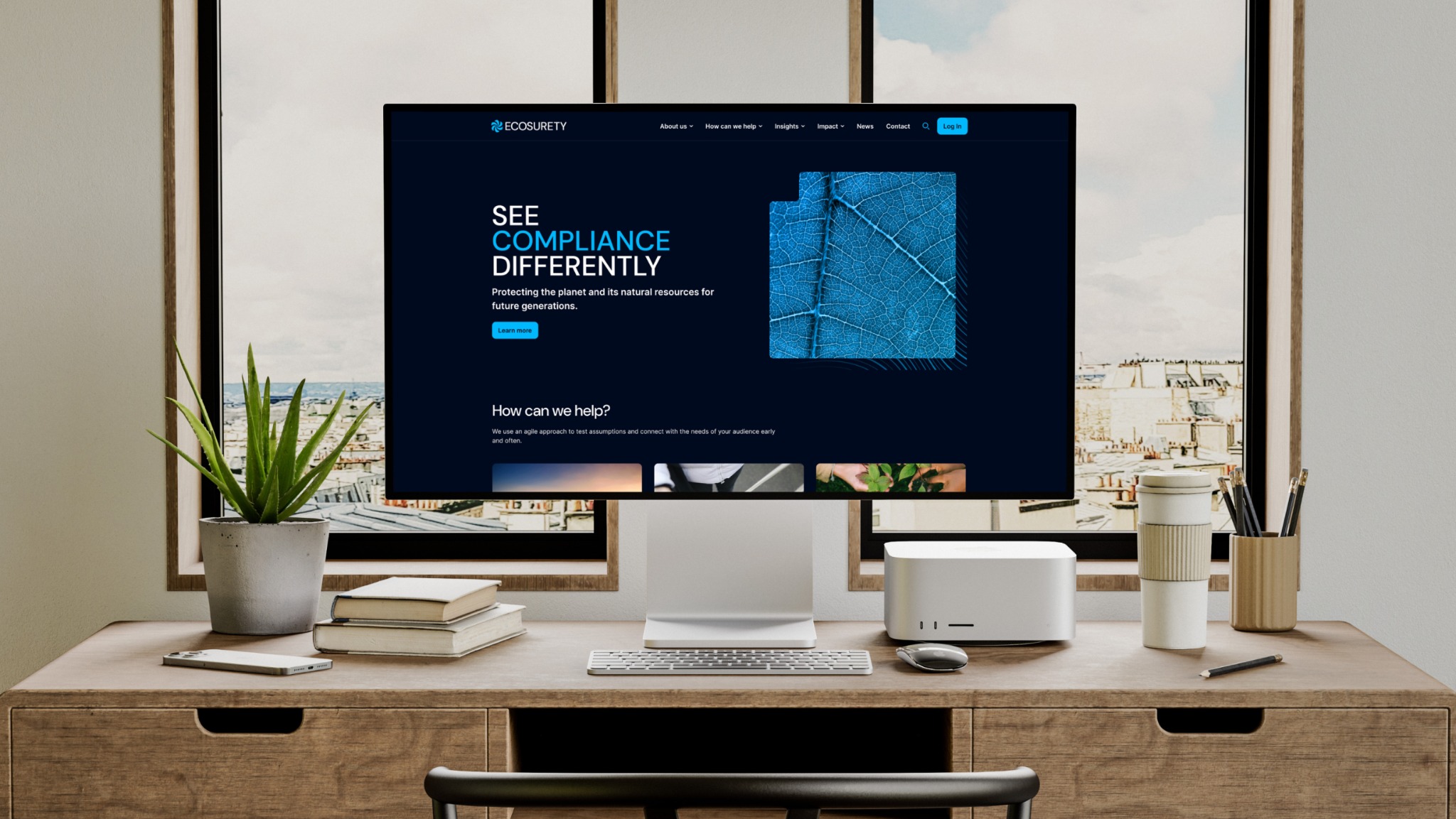 Ekran komputera stacjonarnego wyświetla stronę internetową Ecosurety z hasłem 'See Compliance Differently' i grafiką liścia na ciemnoniebieskim tle, umieszczony na drewnianym biurku z rośliną...