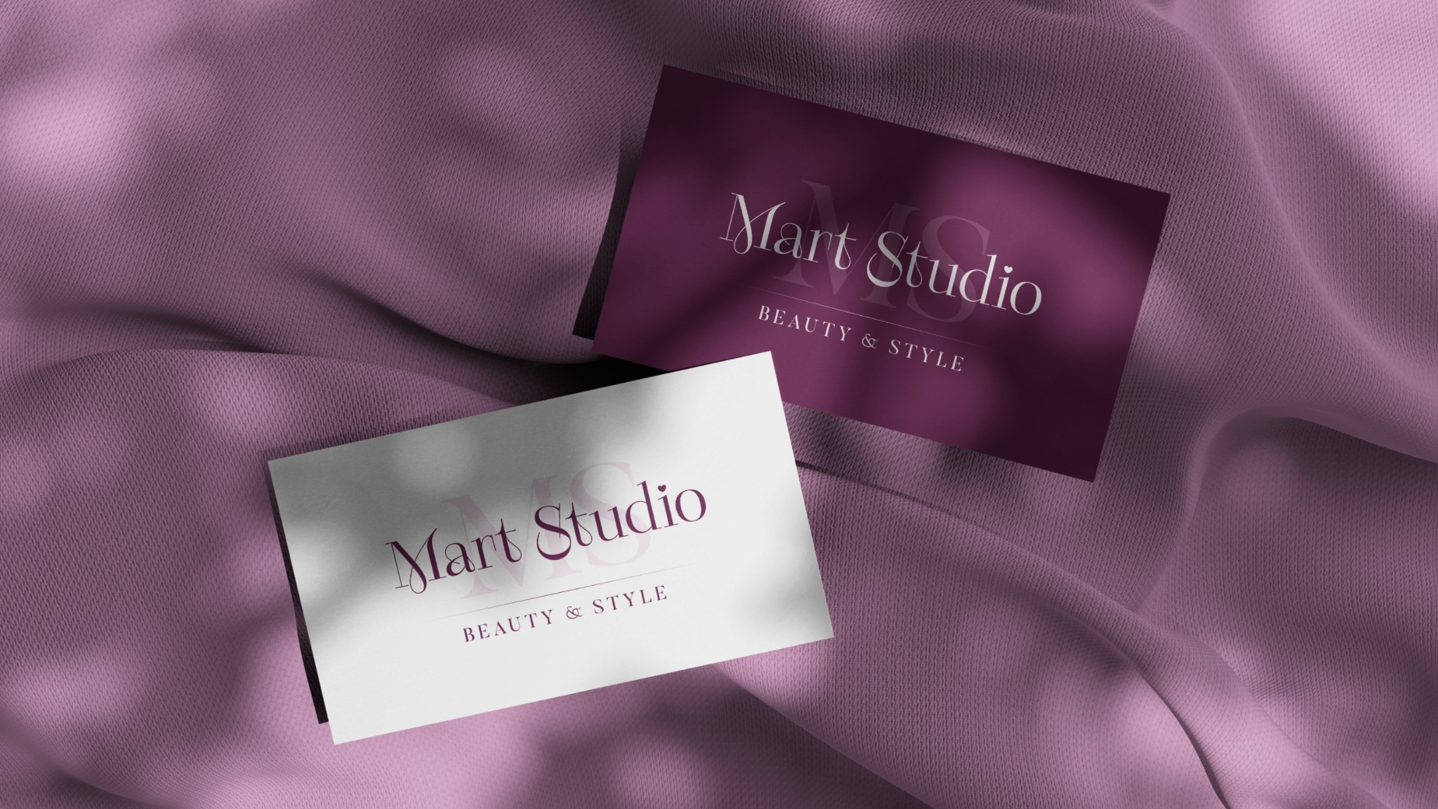 Dwie wizytówki Mart Studio Beauty & Style, jedna biała, druga w kolorze śliwkowym, leżą na fioletowej tkaninie w cieniu liści.