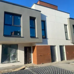 Nowoczesne, szeregowe domy w trakcie budowy, z oknami zabezpieczonymi folią, częściowo wykończoną elewacją w odcieniach szarości, bieli i brązu, widoczne przyłącza instalacyjne.