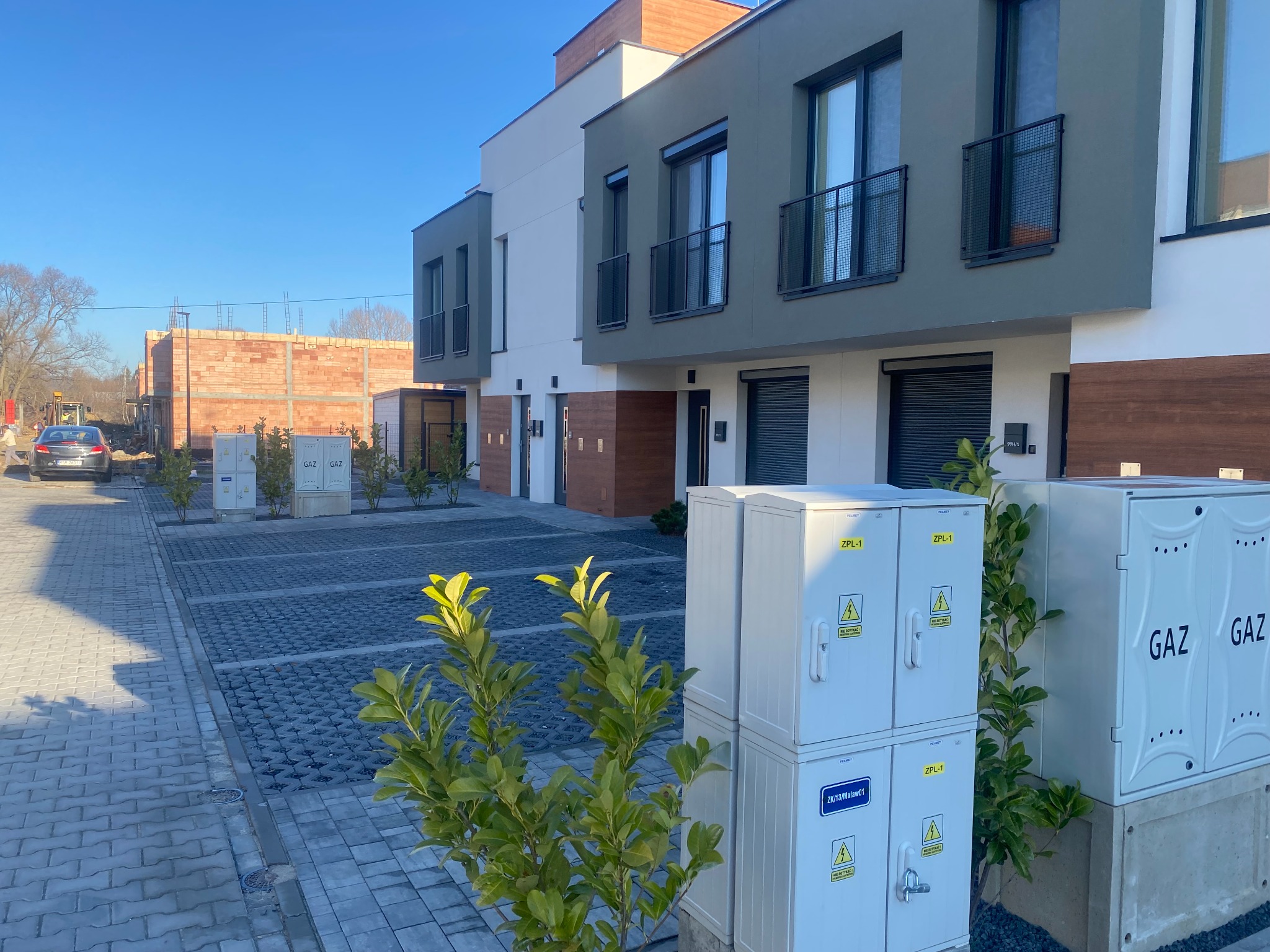 Nowoczesne szeregowe domy z elewacją w odcieniach szarości i bieli, widoczne skrzynki gazowe i elektryczne oraz budynek w trakcie budowy w tle.