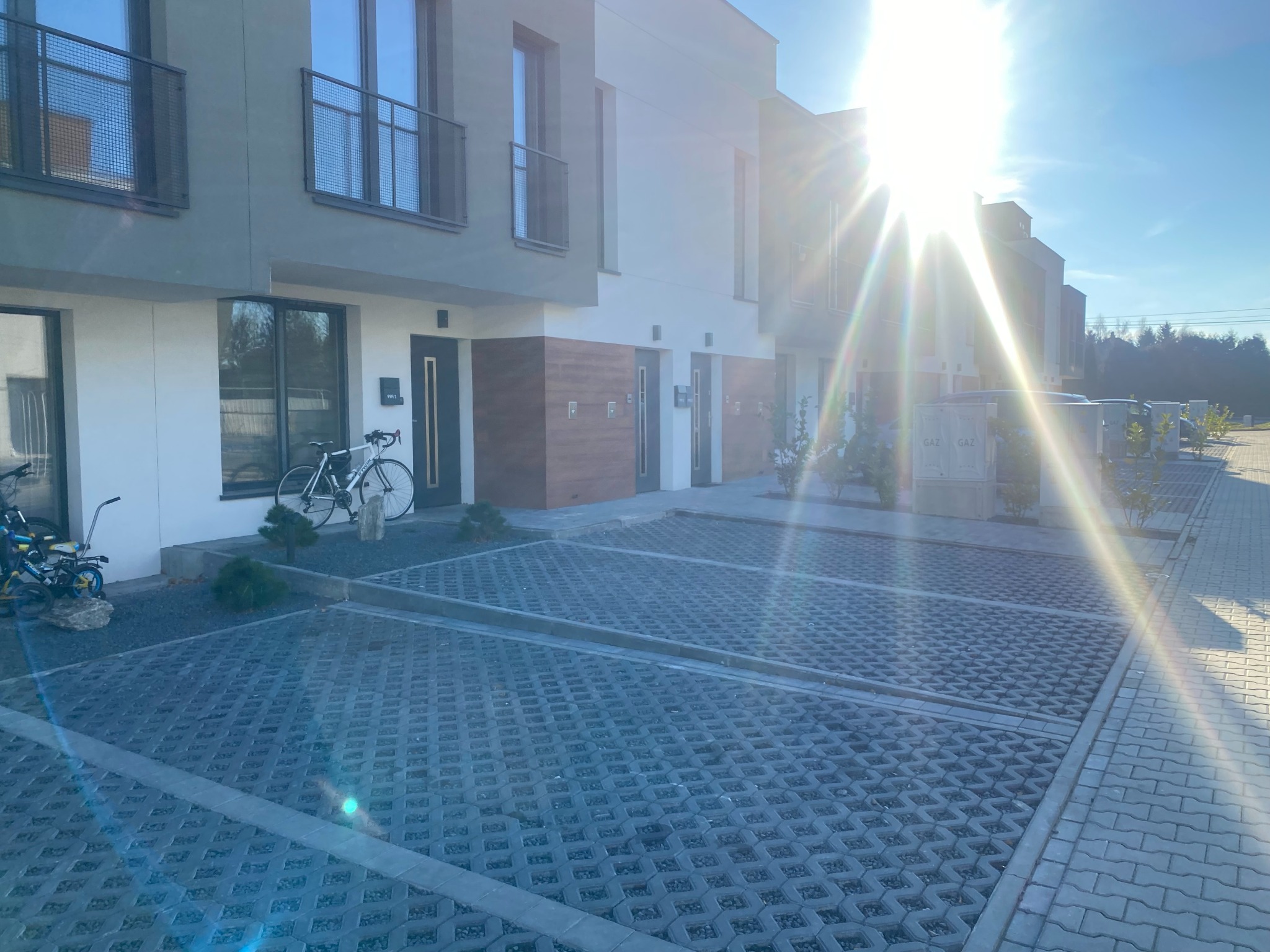 Rząd nowo wybudowanych domów jednorodzinnych z szarymi elewacjami, widoczne rowery przed jednym z nich i ażurowy parking z kostki betonowej w słoneczny dzień.