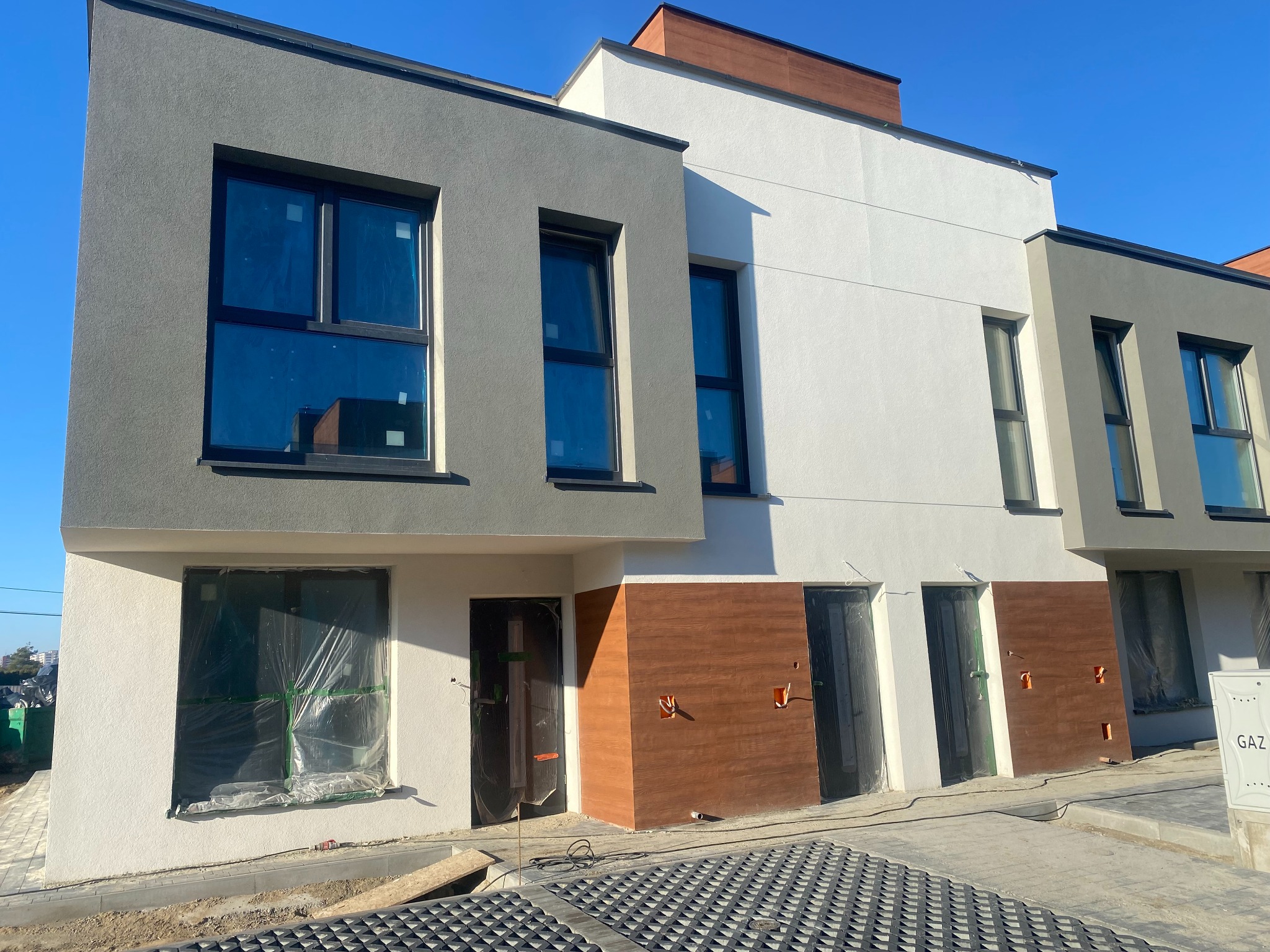 Nowoczesne, szeregowe domy w trakcie budowy, z oknami zabezpieczonymi folią, częściowo wykończoną elewacją w odcieniach szarości, bieli i brązu, widoczne przyłącza instalacyjne.