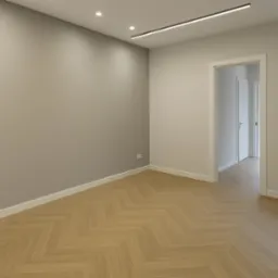 Puste, jasne pomieszczenie z parkietem ułożonym w jodełkę, szarą ścianą, białymi listwami przypodłogowymi i nowoczesnym oświetleniem sufitowym.