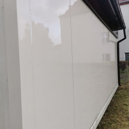 GlassCare - Boczny widok wyczyszczonej, błyszczącej elewacji budynku z jasnymi panelami, z widoczną rynną i fragmentem trawnika. Świeżo umyta powierzchnia odbija niebo.