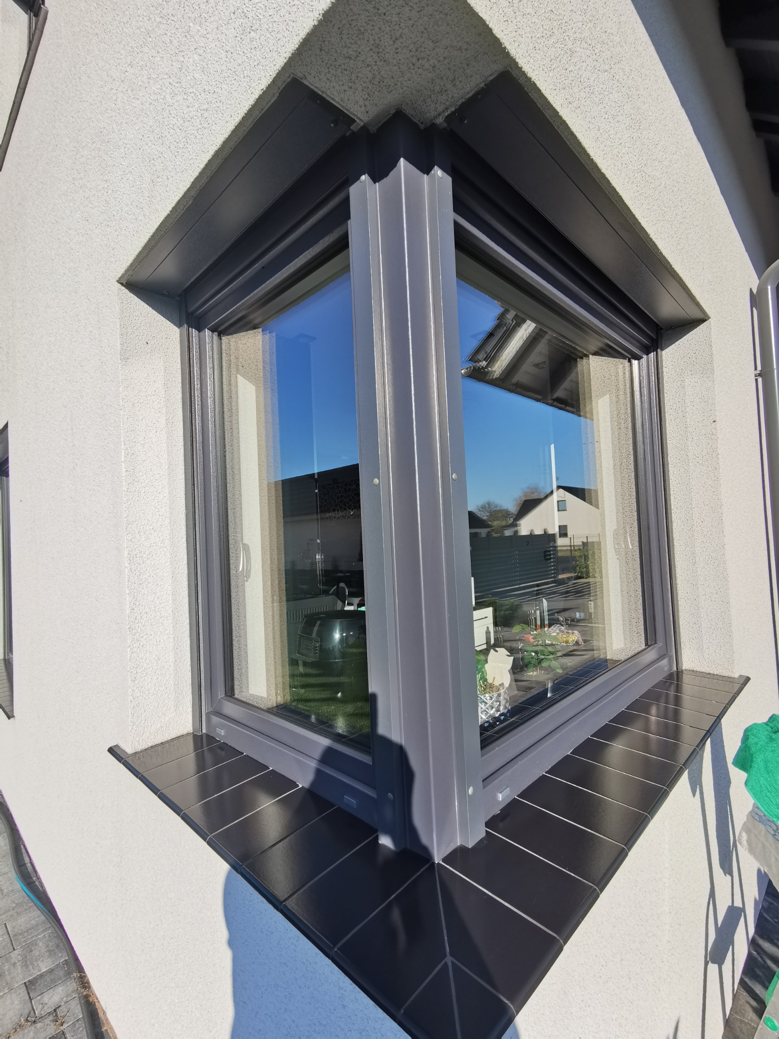 Szare okno narożne z czarnym parapetem z płytek, odbijające błękit nieba i dom. Widoczny fragment elewacji z fakturą baranka i zielona ściereczka.