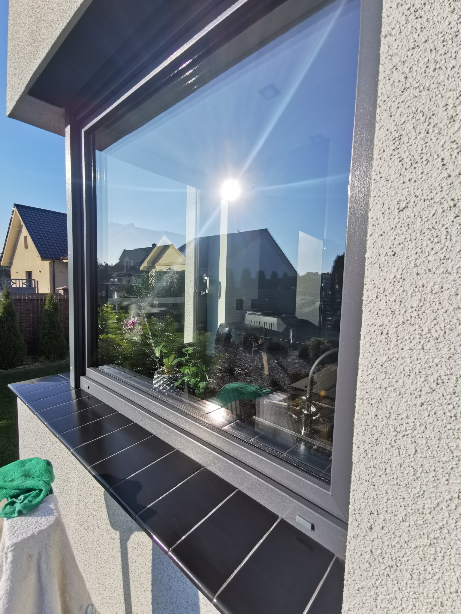 Błyszczące, umyte okno z widokiem na ogród i domy w słoneczny dzień. Na parapecie czarne płytki i zielona ściereczka. Widoczne refleksy słońca na szybie.