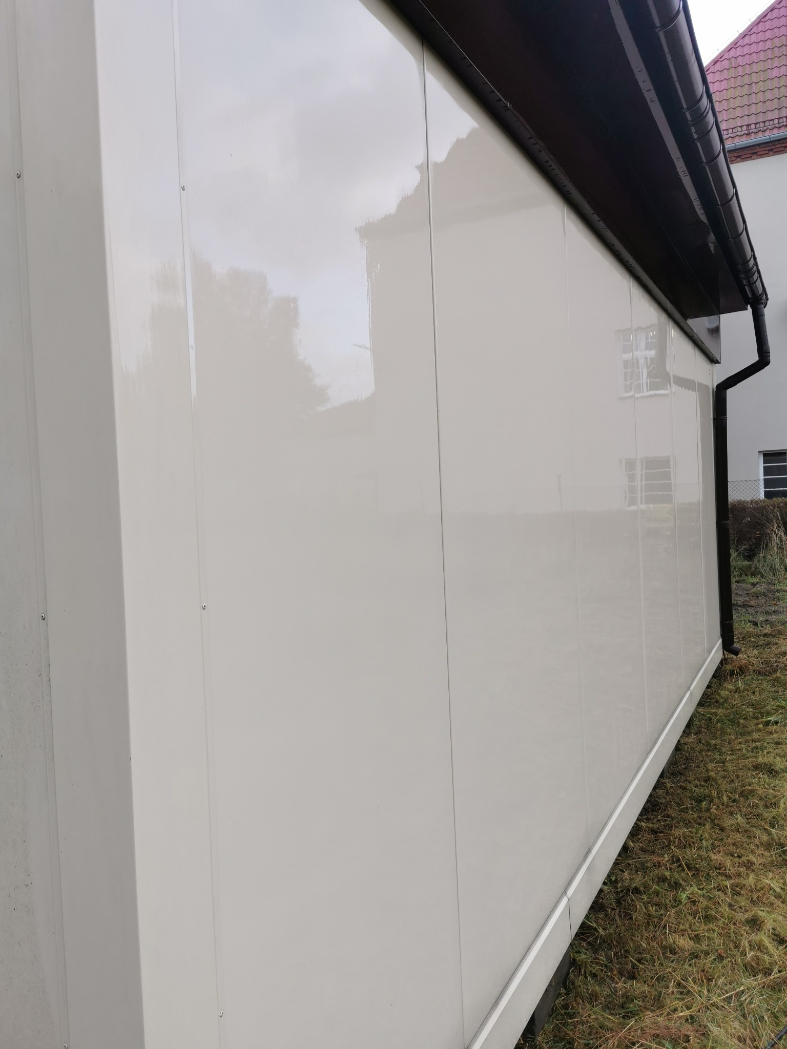Boczny widok wyczyszczonej, błyszczącej elewacji budynku z jasnymi panelami, z widoczną rynną i fragmentem trawnika. Świeżo umyta powierzchnia odbija niebo.