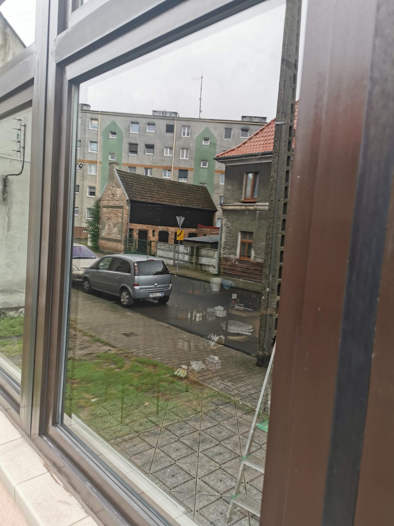 Widok z okna na ulicę z zaparkowanymi autami i budynkami w tle. W rogu widoczna drabina oparta o ścianę. Okno z wyraźnymi refleksami.