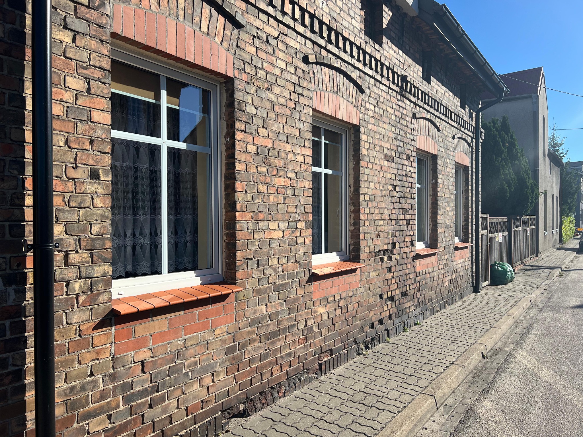 Murek z cegły klinkierowej z trzema oknami PCV. Widoczne zabrudzenia na murze, chodnik z kostki brukowej i ulica. Słoneczny dzień.