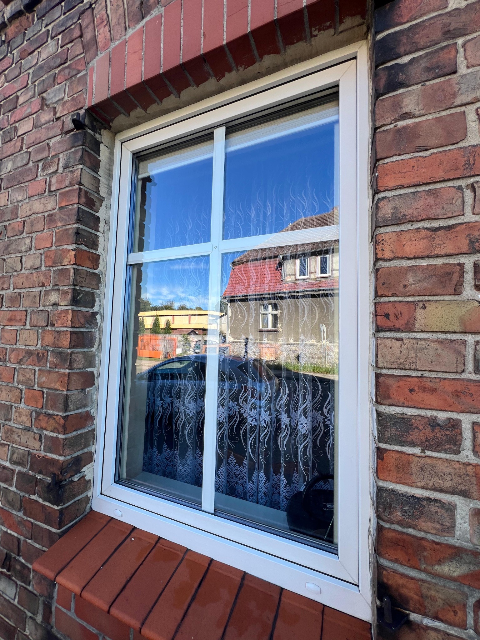 Białe okno z szybami dzielonymi na cztery części, w czerwonej ceglanej ścianie. W szybie odbija się błękitne niebo i budynek z czerwonym dachem.