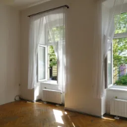 Jasne wnętrze z drewnianą podłogą, dwoma oknami z białymi firankami i grzejnikami. Komoda z szufladami w rogu. Widok na zieleń za oknami.