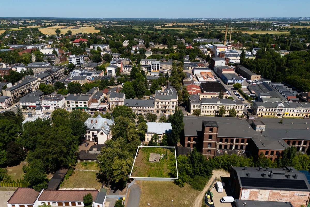 Widok z lotu ptaka na działkę w mieście, obrysowaną białą linią. W tle zabudowa miejska i zieleń. Działka z pozostałościami fundamentów.