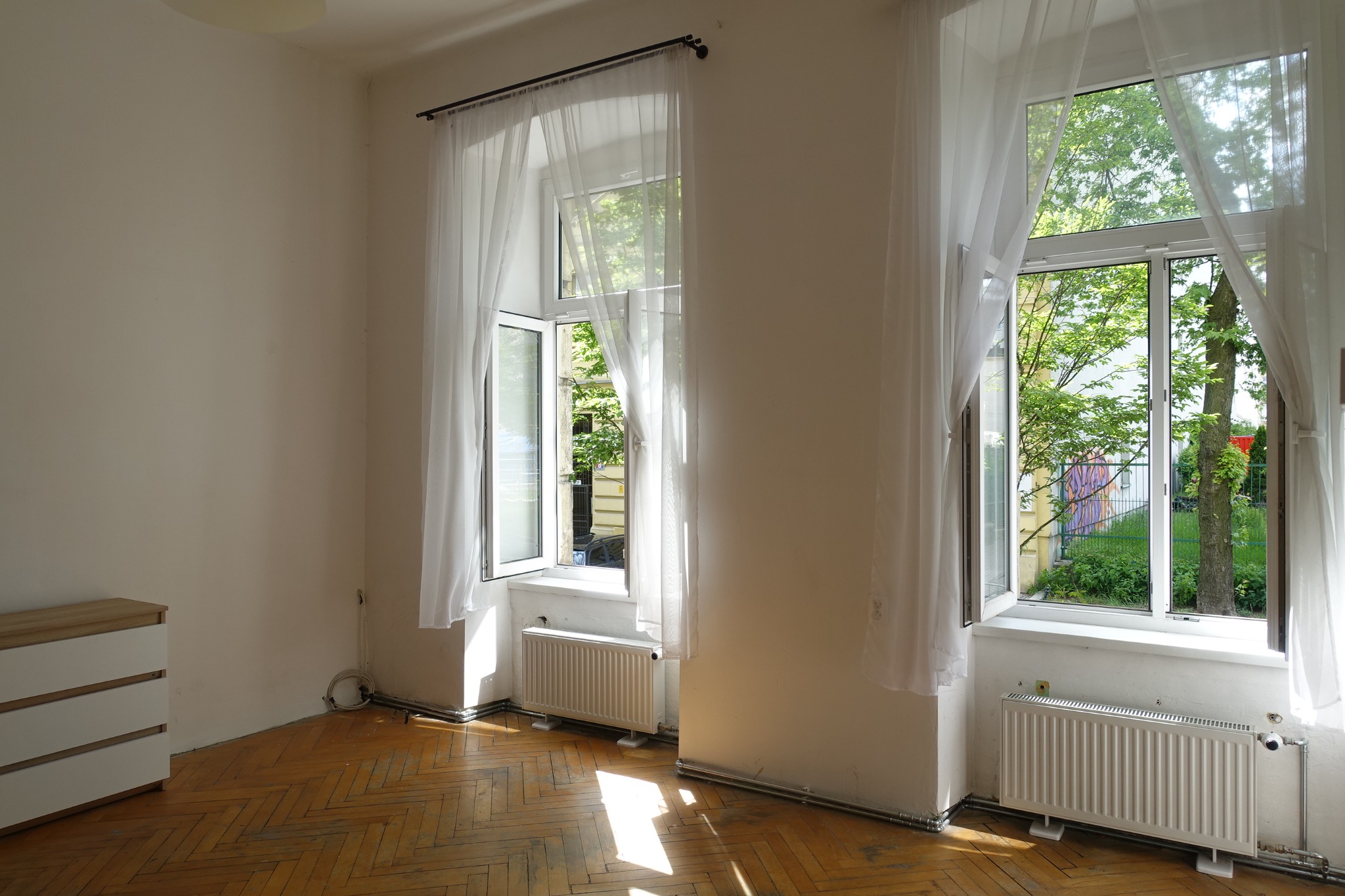 Jasne wnętrze z drewnianą podłogą, dwoma oknami z białymi firankami i grzejnikami. Komoda z szufladami w rogu. Widok na zieleń za oknami.
