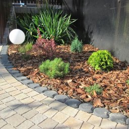 PERSSON PRZEMYSŁAW JABŁOŃSKI - Rabata z różnorodnymi krzewami iglastymi i liściastymi, wysypana korą, obrzeżona kamienną ścieżką, z kulistą lampą ogrodową w tle schodów.