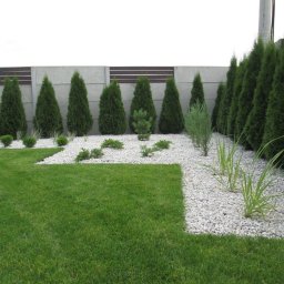 PERSSON PRZEMYSŁAW JABŁOŃSKI - Starannie zaprojektowany ogród z równo przystrzyżonym trawnikiem, białymi kamykami, rzędem tuj i niskimi krzewami, oddzielony od sąsiedniej posesji betonowym ogrodzeniem.