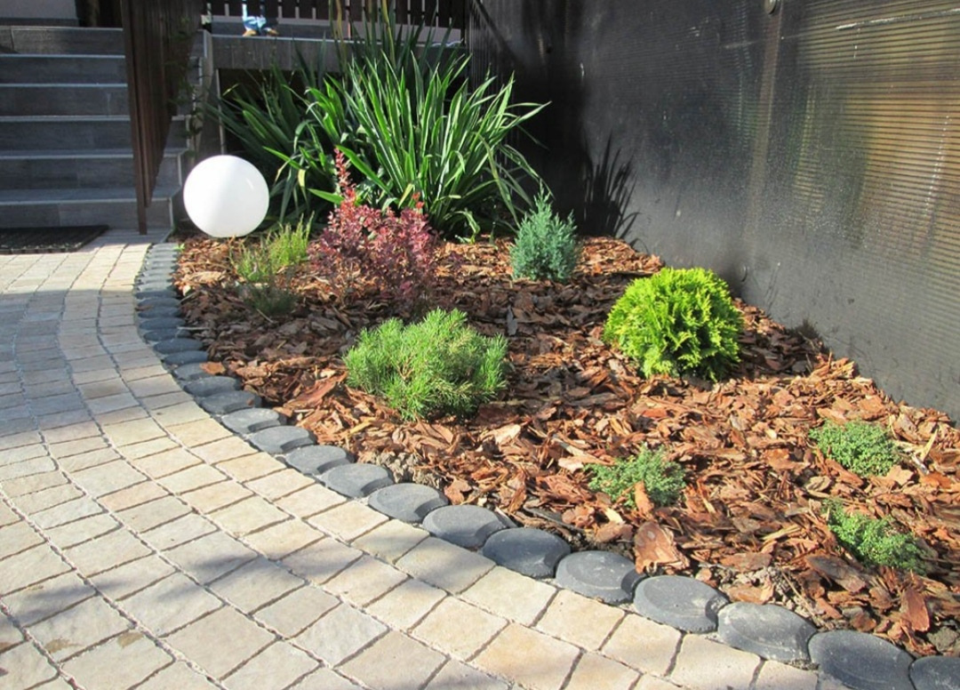 Rabata z różnorodnymi krzewami iglastymi i liściastymi, wysypana korą, obrzeżona kamienną ścieżką, z kulistą lampą ogrodową w tle schodów.