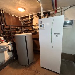 Artur Górczyński Pema - Biała pompa ciepła Ecodan z szarym zbiornikiem buforowym w kotłowni. Widoczne rury instalacji grzewczej. Oświetlenie sufitowe, brązowe ściany i płytki podłogowe.