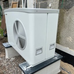 Artur Górczyński Pema - Biała pompa ciepła Stiebel Eltron zamontowana na betonowych podstawach przy budynku z ocieploną elewacją i folią kubełkową. Widoczne elementy instalacyjne.