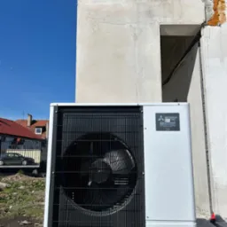 Biała pompa ciepła Mitsubishi Zubadan R32 na zewnątrz budynku w trakcie budowy, widoczna instalacja, błękitne niebo. Ujęcie z dołu podkreśla wielkość urządzenia.