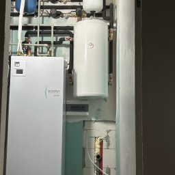 Artur Górczyński Pema - Biała pompa ciepła Ecodan z zbiornikiem i instalacją w surowym wnętrzu. Widoczne rury, zawory i fragment nieotynkowanej ściany z instalacją.