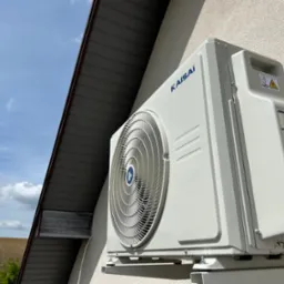 Biała jednostka zewnętrzna klimatyzacji Kaisai zamontowana na ścianie budynku, widok z dołu, z fragmentem dachu i błękitnym niebem w tle.