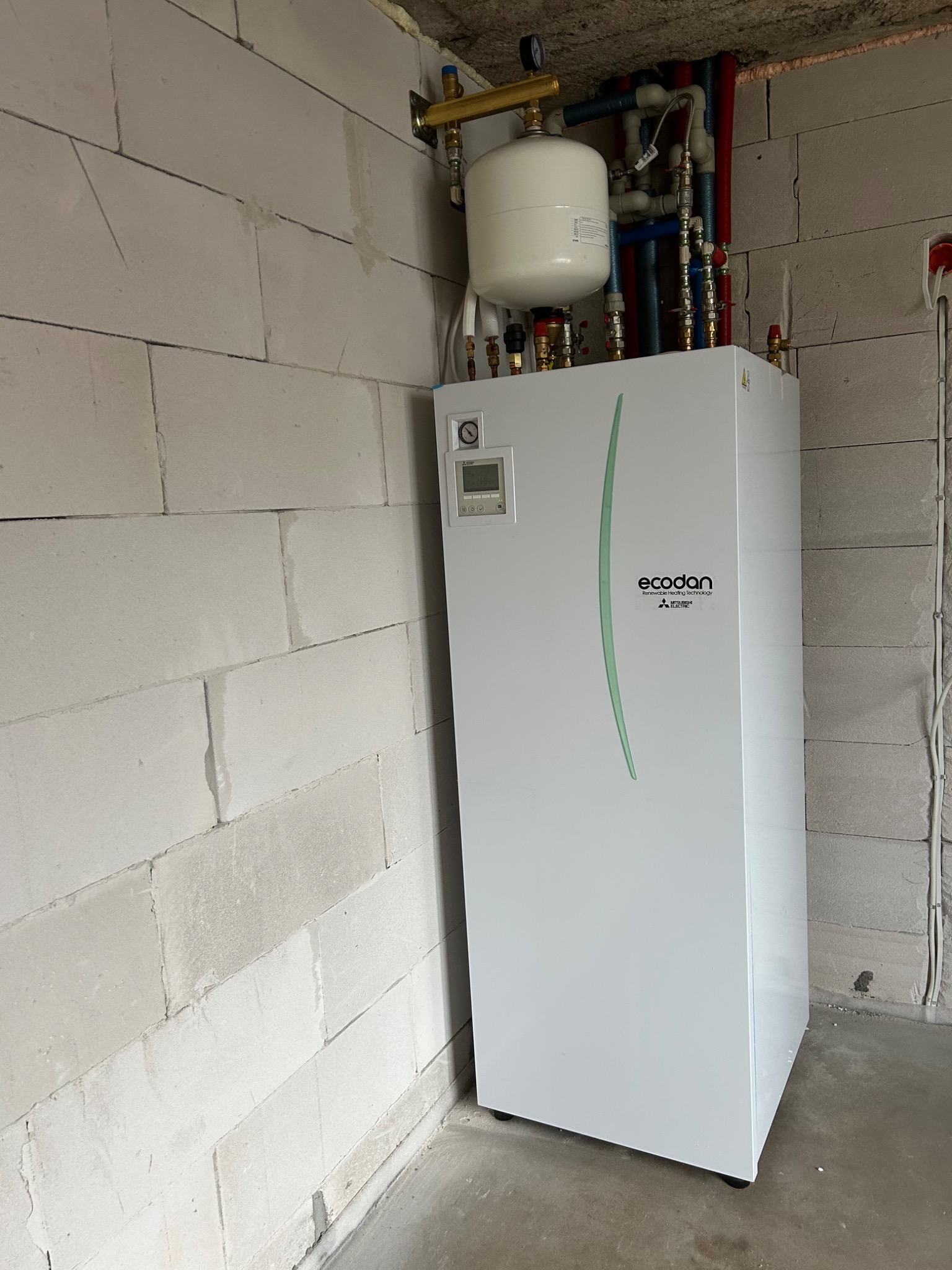 Biała pompa ciepła Ecodan Mitsubishi Electric z zbiornikiem wyrównawczym na tle ściany z bloczków betonu komórkowego w surowym stanie.