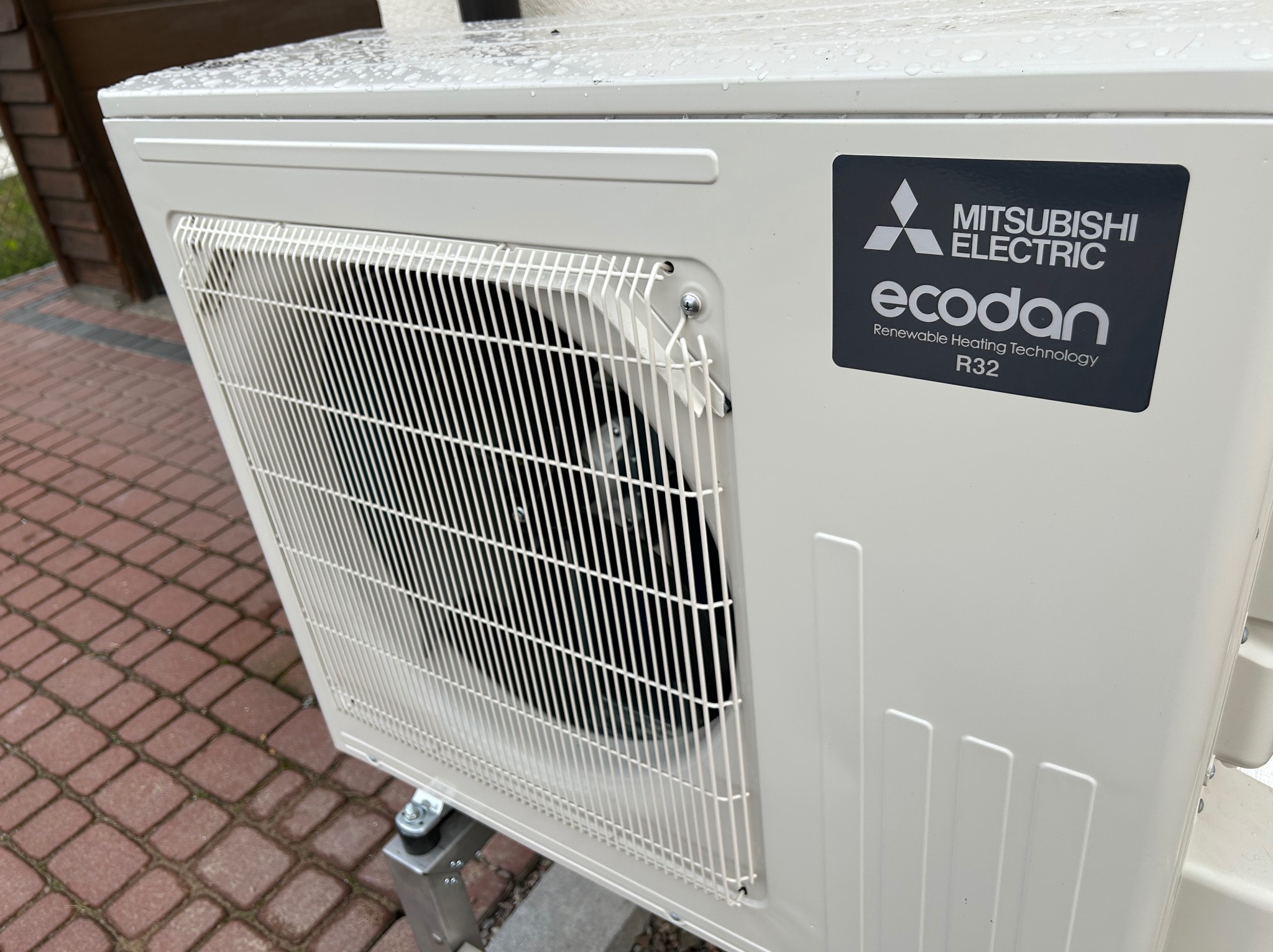 Biała, zewnętrzna jednostka pompy ciepła Mitsubishi Ecodan R32 na kostce brukowej, widoczna z boku, z bliska. Na obudowie naklejka z logo producenta.