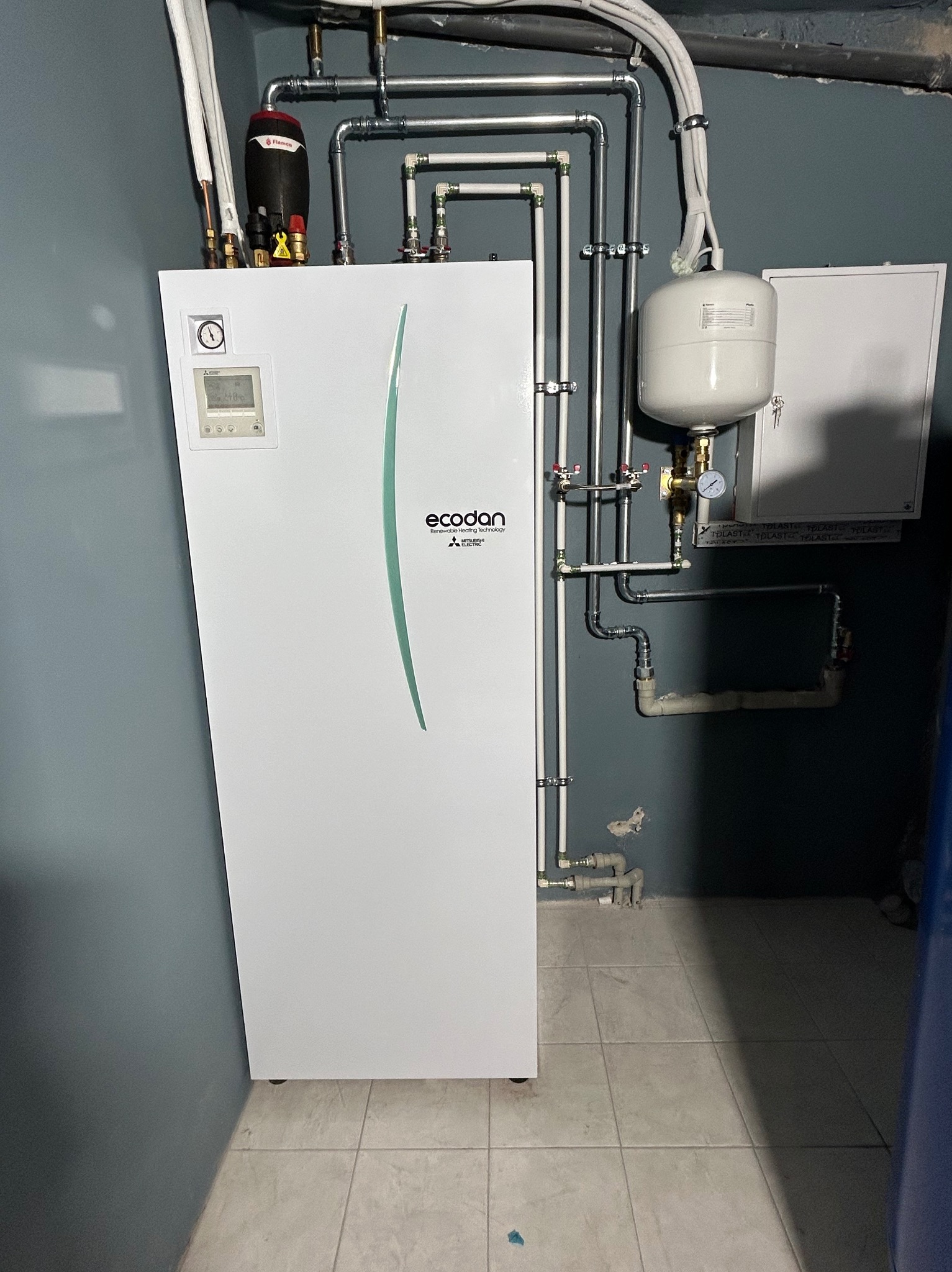 Biała pompa ciepła Ecodan Mitsubishi z szarą, pionową aplikacją, widoczne rury instalacji grzewczej na tle ciemnoszarej ściany i jasnej podłogi z płytek.