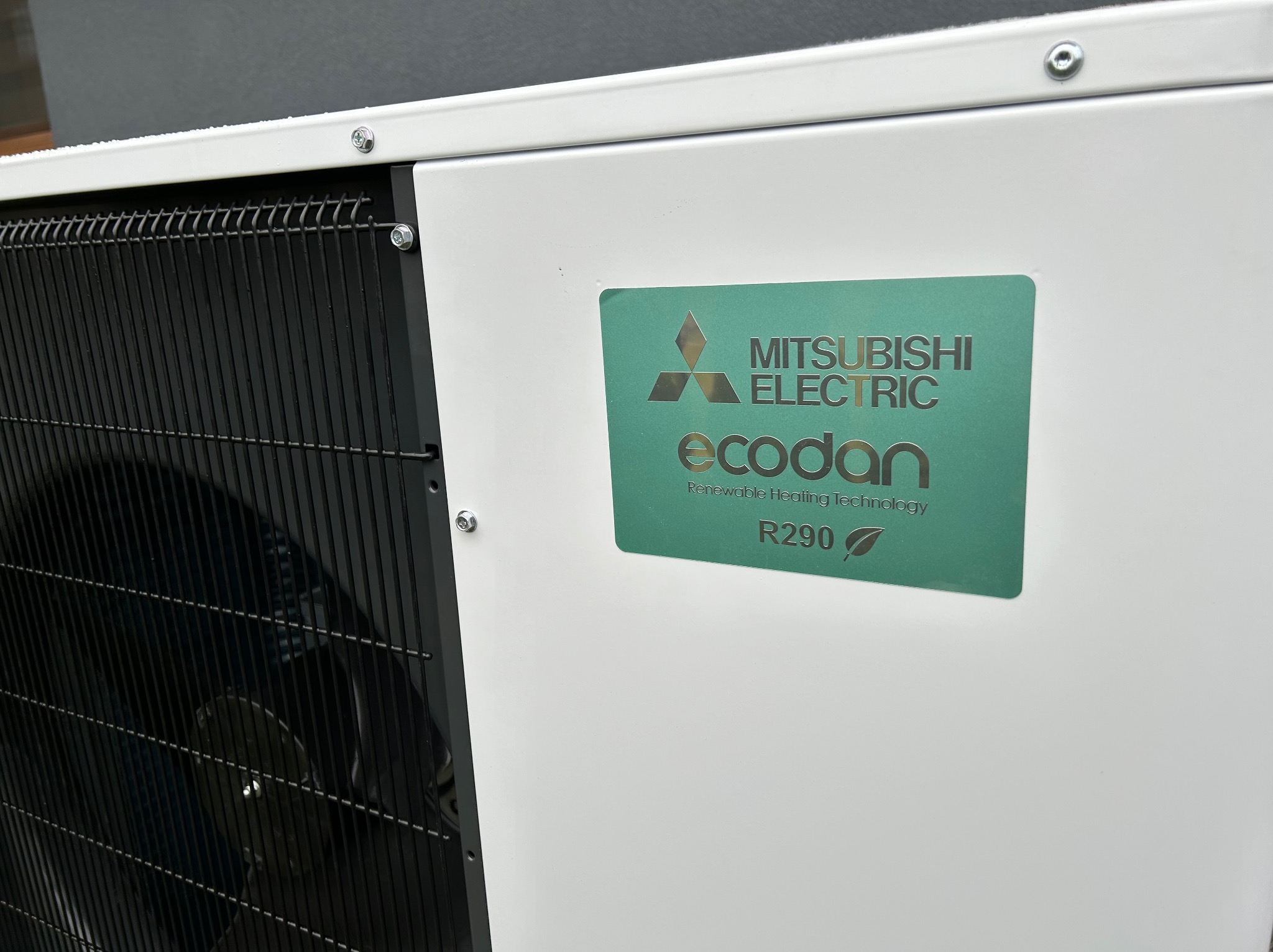 Biała pompa ciepła Mitsubishi Electric Ecodan R290 z widoczną czarną kratką wentylacyjną, zamontowana na zewnątrz budynku.