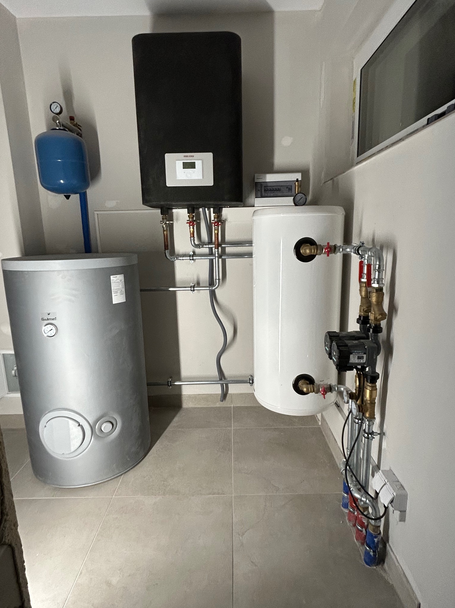 Zestaw instalacji grzewczej: czarny piec z panelem, biały i srebrny zbiornik, niebieski zbiornik ciśnieniowy, rury miedziane i zawory w schludnym, jasnym pomieszczeniu.