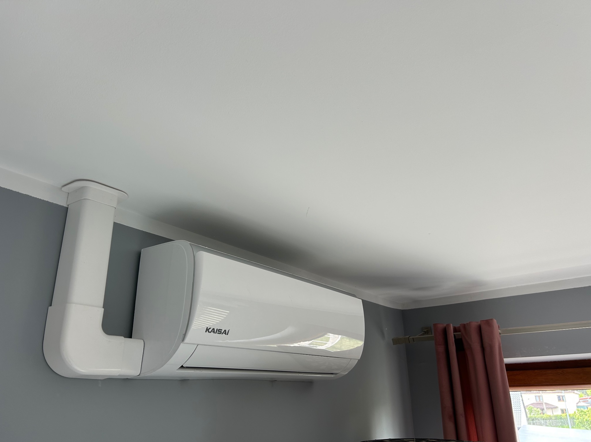 Biały klimatyzator Kaisai zamontowany na szarej ścianie, z widoczną instalacją prowadzącą do sufitu. Fragment okna z zasłoną w tle.