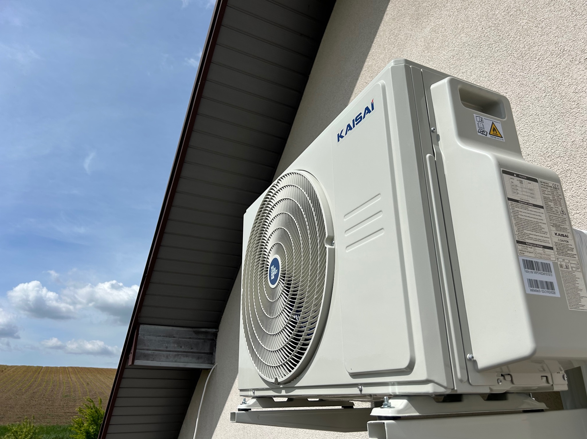 Biała jednostka zewnętrzna klimatyzacji Kaisai zamontowana na ścianie budynku, widok z dołu, z fragmentem dachu i błękitnym niebem w tle.