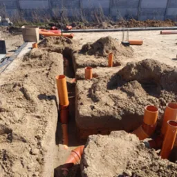 Wykonane wykopy pod instalacje sanitarne z ułożonymi pomarańczowymi rurami kanalizacyjnymi, widoczne złącza i kolanka, teren budowy z piaskiem, w tle betonowe ogrodzenie.