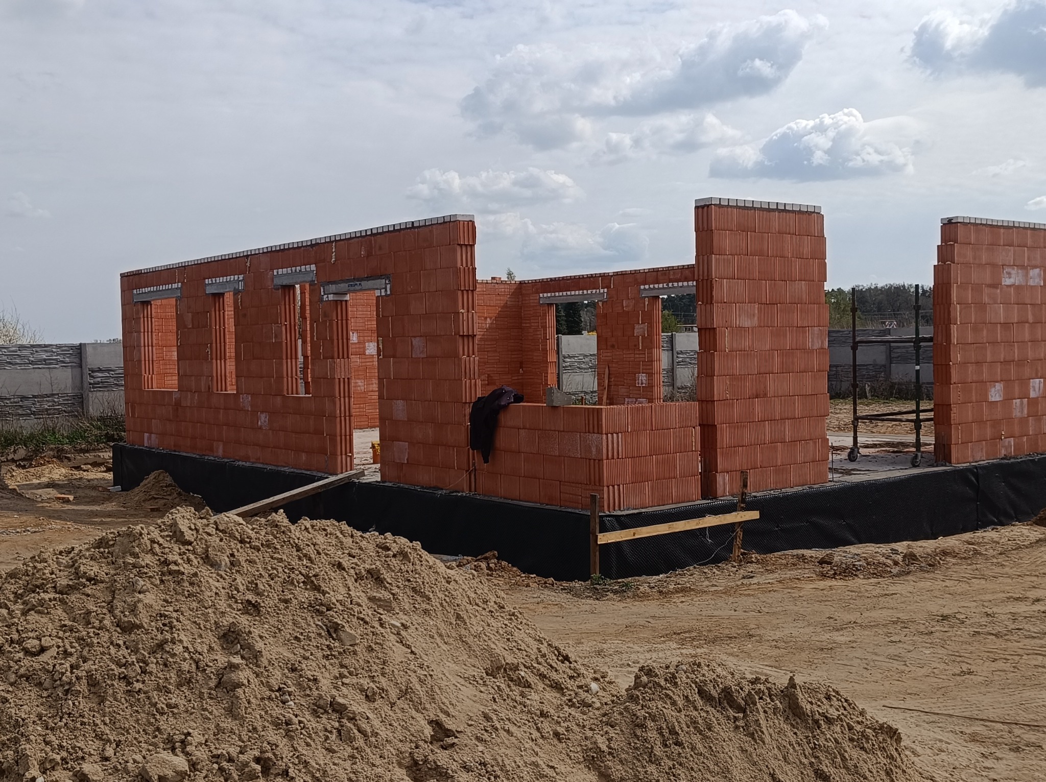Wznoszenie ścian domu z czerwonej cegły ceramicznej na fundamencie zabezpieczonym czarną folią hydroizolacyjną, widoczne otwory okienne i stropy, hałda piasku na pierwszym planie.