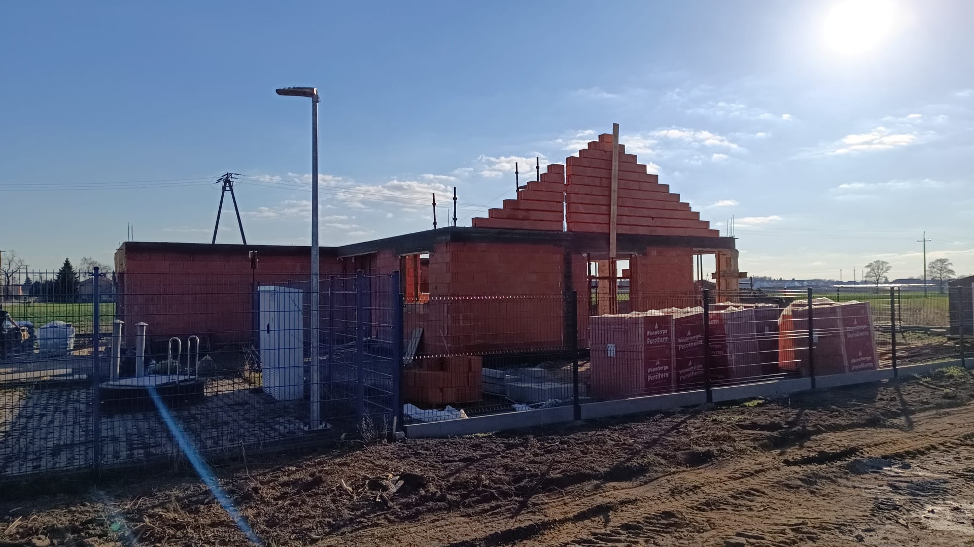 Wznoszenie ścian domu z czerwonej cegły Porotherm, widoczne palety z cegłami, ogrodzenie posesji z siatki, słoneczny dzień.