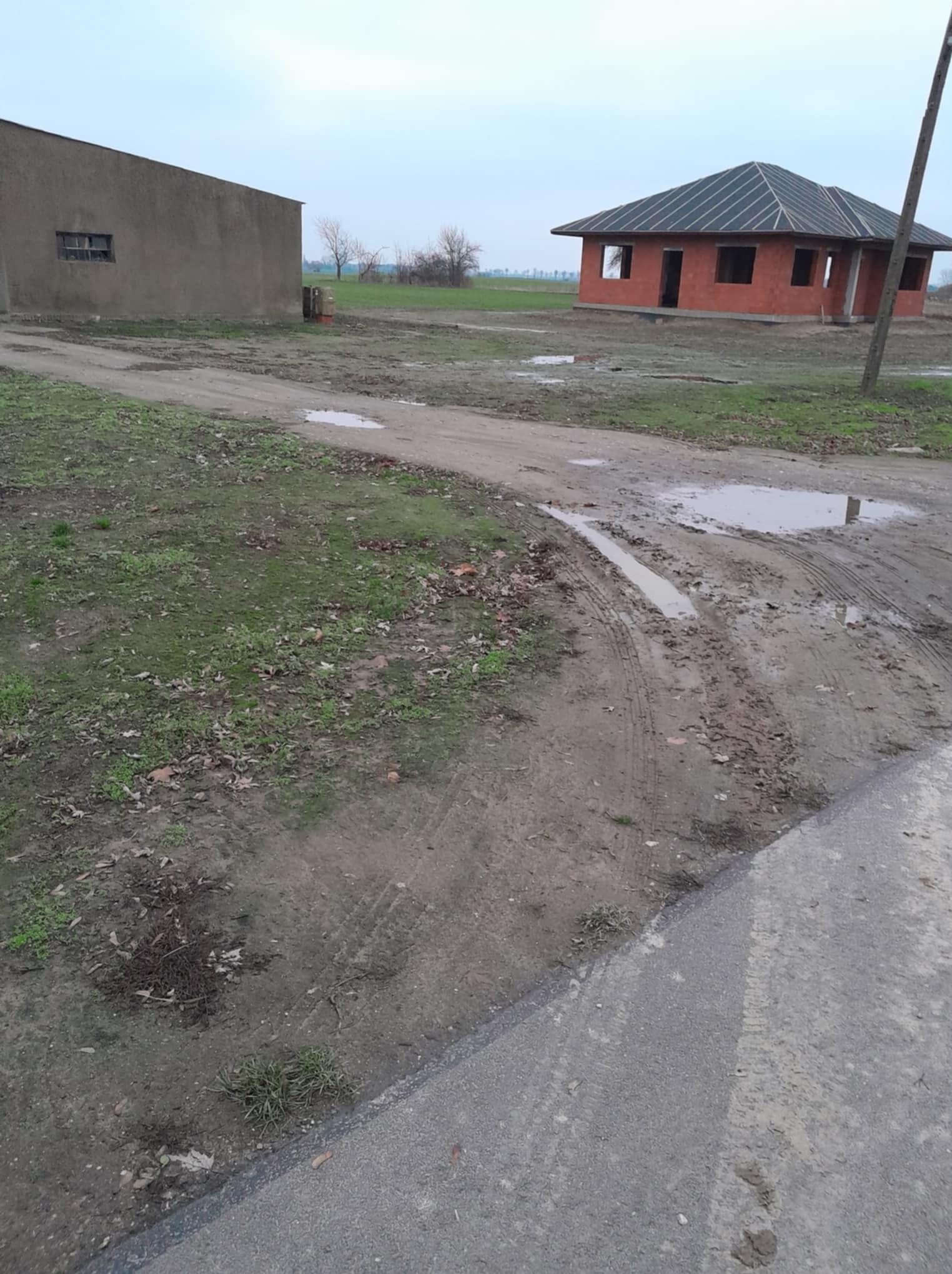 Niedokończony budynek z czerwonej cegły z dachem z dachówki bitumicznej na błotnistym terenie, widoczne ślady opon i kałuże.