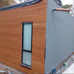 Budynek z elewacją częściowo wykończoną drewnianą okładziną i szarą strukturą, widoczne rury odprowadzające wodę i okno z czarną ramą, teren budowy.