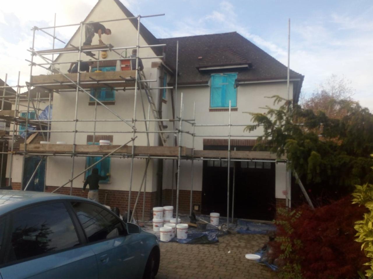 Dom jednorodzinny w trakcie malowania elewacji, okna zabezpieczone folią, rusztowania rozstawione, widoczne puszki z farbą i zaparkowany samochód.