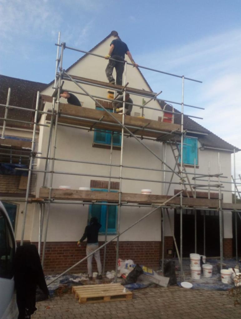 Prace na elewacji domu jednorodzinnego, widoczne rusztowania, pracownicy w trakcie wykonywania prac, okna zabezpieczone folią, na ziemi wiadra z farbą i paleta.