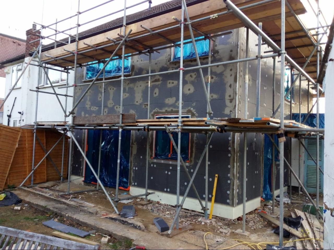 Dom w trakcie ocieplania, pokryty czarnymi płytami izolacyjnymi, zabezpieczone okna folią, rusztowanie wokół budynku.